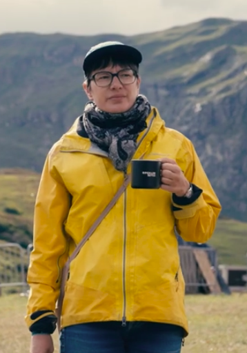 A handsome lady with a yellow Regenjacke trying to look intellectual in front of a mountain landscape.